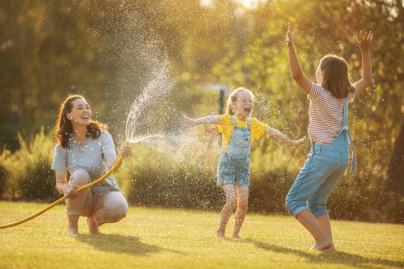 A mom crouching with a garden hose and spraying her daughters with water as they all laugh and the girls play in the water.