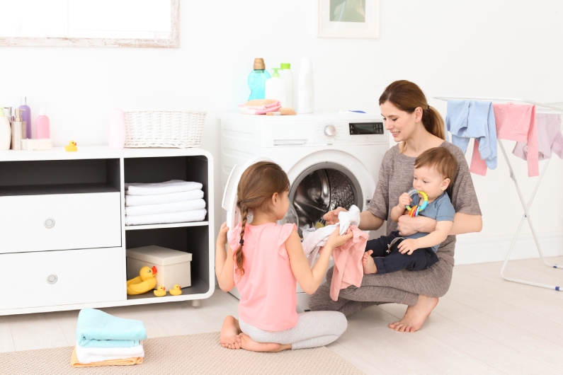 A mom and her two young toddlers working on household chores together. They're taking clothes out of the dryer.