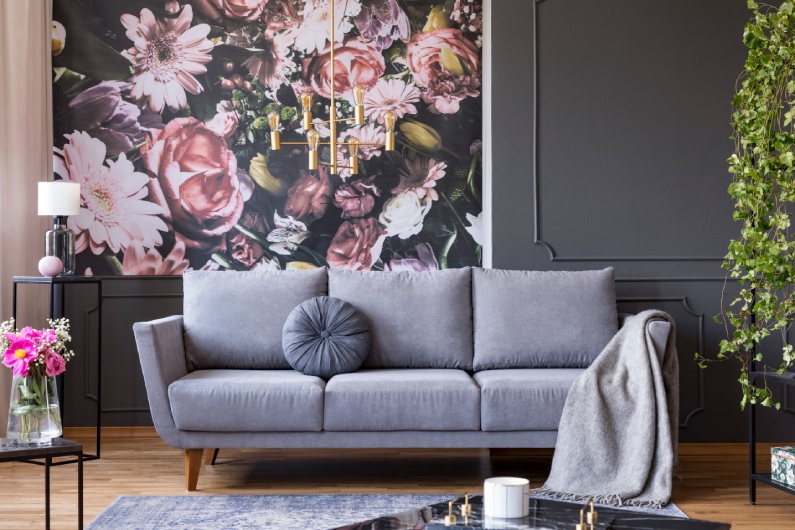 A contemporary living room with dark floral wallpaper, a gray couch with a throw pillow and blanket, and matching tables.