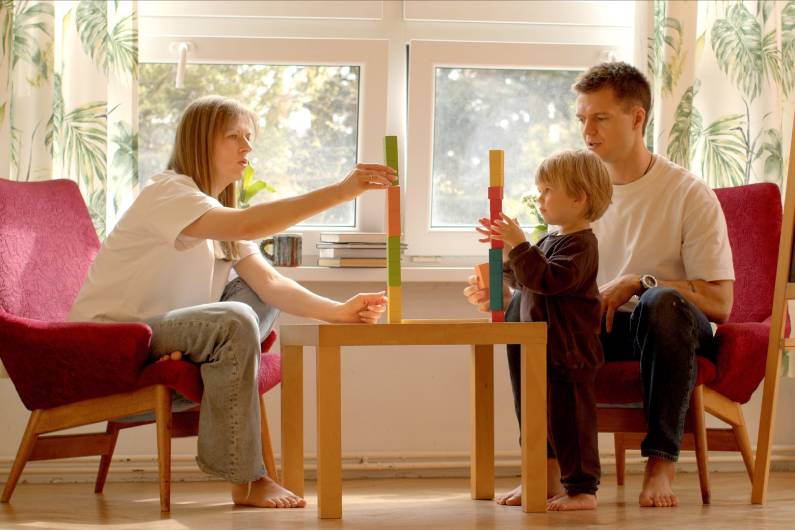 Parents bonding with their young child as they all play and build blocks. The blocks are colorful and the child is standing.