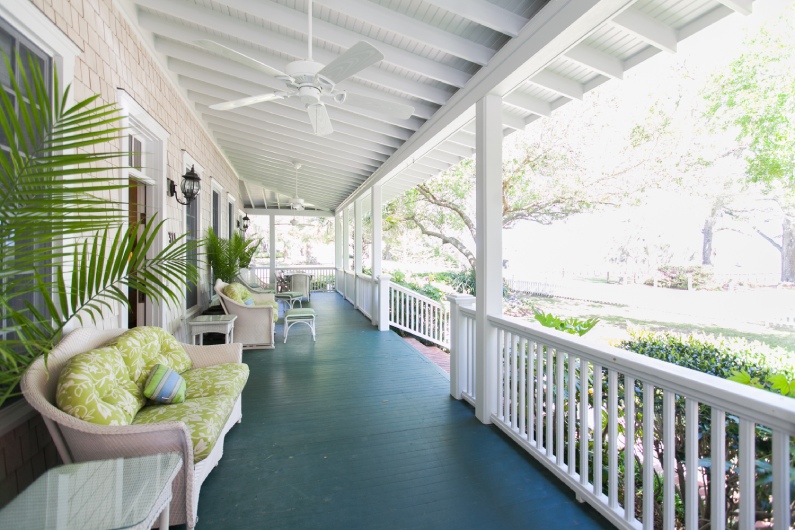 A long wrap-around porch with white railings, green wood floors, wicker furniture, and potted palm plants.