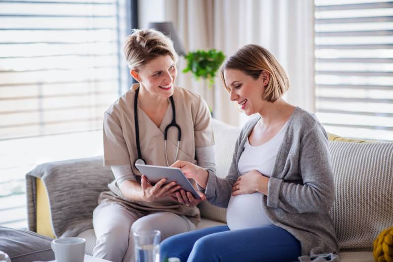 A woman in scrubs and a pregnant woman sitting on a couch, smiling, and referencing a tablet between them.