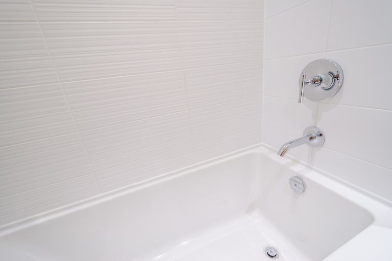 A residential bathroom with a plain white bathtub surround. The bathtub and shower have stainless steel fixtures.