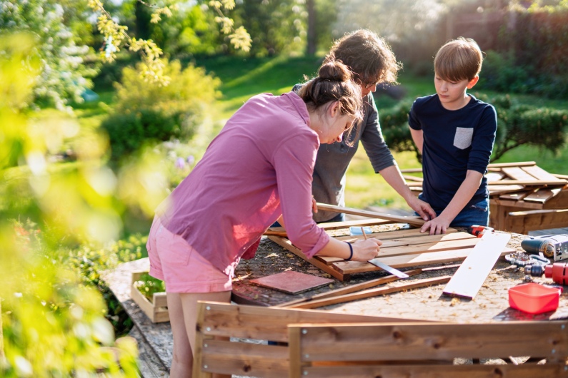 A woman and two male children outside on a sunny day surrounded by trees as they work on building something with wood.