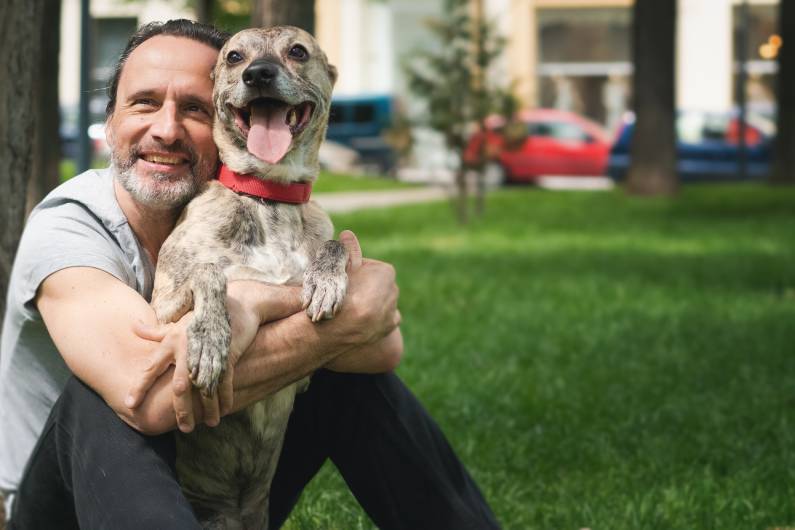 A smiling man sits in the grass, hugging a dog. The dog is standing on its hind legs, mouth open and tongue sticking out.