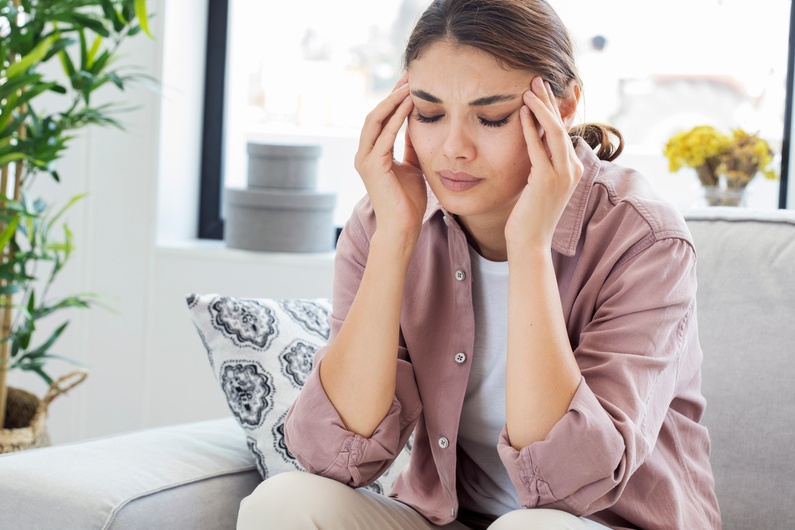 A woman in a button up shirt sits on the couch and presses her fingers into her temples to alleviate a headache.